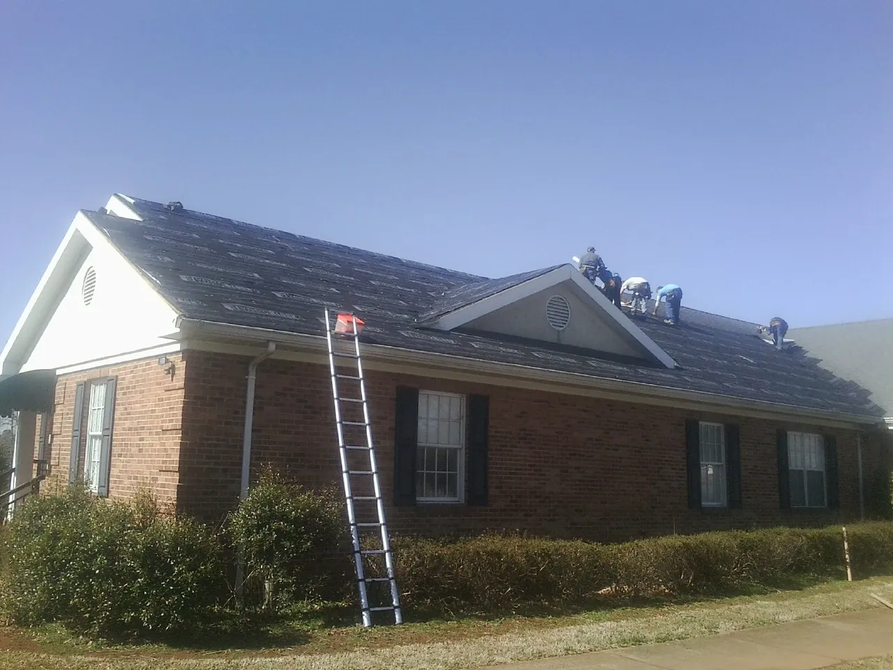 Crew tearing shingles off a brick ranch roof with a ladder set — Emergency Roof Repair work by our crew in High Springs